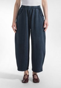 Osoba ubrana w luźne, ciemnoniebieskie spodnie z elastycznym paskiem w talii i bocznymi kieszeniami oraz czerwone, sznurowane buty na płaskim obcasie na białym tle.