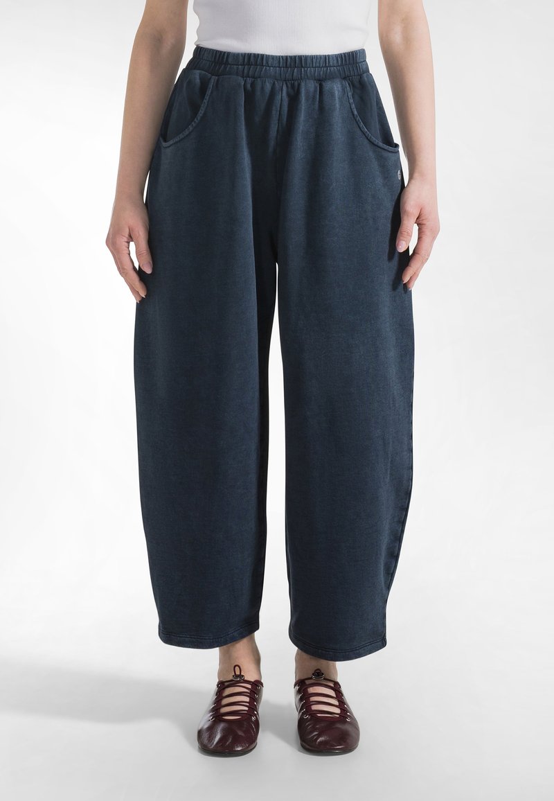 Osoba ubrana w luźne, ciemnoniebieskie spodnie z elastycznym paskiem w talii i bocznymi kieszeniami oraz czerwone, sznurowane buty na płaskim obcasie na białym tle.