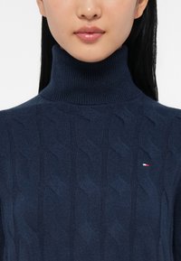 Nainen, jolla on yllään tummansininen palmikkoneulepusero, jossa on pieni punainen, valkoinen ja sininen logo rinnassa.