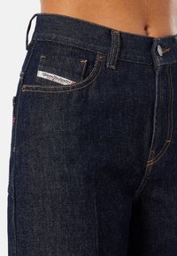 Detailní záběr na pas a horní část stehna tmavě modrých džínů s viditelnou nášivkou "Diesel Industry Denim Division".