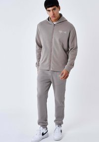 Sweat à capuche gris zippé avec cordon de serrage, au design épuré, poches avant, et joggeurs gris assortis avec accents de logo. Baskets blanches.
