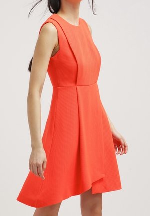 Kvinde iført en ærmeløs, lys orange tekstureret kjole med asymmetrisk kant mod en ensfarvet lys baggrund.