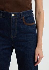 Jeans de mezclilla azul oscuro con una cintura alta, ribetes de bolsillo en marrón, cierre de botón dorado y costuras amarillas en contraste.