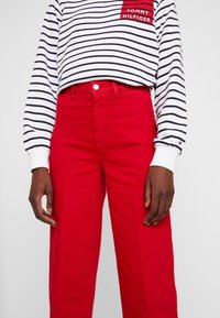 Persona che indossa pantaloni rossi e una maglietta a maniche lunghe bianca con righe nere e una toppa rossa del logo Tommy Hilfiger.
