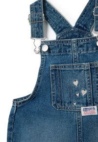 Denim-Latzhose in mittlerem Waschzustand mit silbernem Zubehör. Sie verfügt über verstellbare Träger, eine Vordertasche mit Herzmustern und einem Etikett.