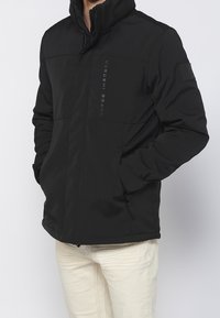 Veste noire isolée avec un col montant et une fermeture éclair. Présente un logo sur le devant et des poches latérales avec fermetures éclair.