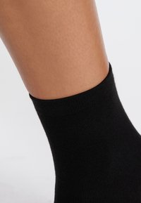 Sort bomuldsstrømpe med ribbet kant, tætsiddende design og glat tekstur, der delvist dækker huden på en ankel.