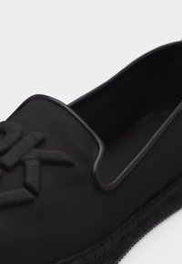Μαύρο παπούτσι τύπου slip-on με υφασμάτινη επάνω επιφάνεια με υφή, ανάγλυφο λογότυπο "CK" στο μπροστινό μέρος και πλεγμένη λεπτομέρεια στη σόλα.