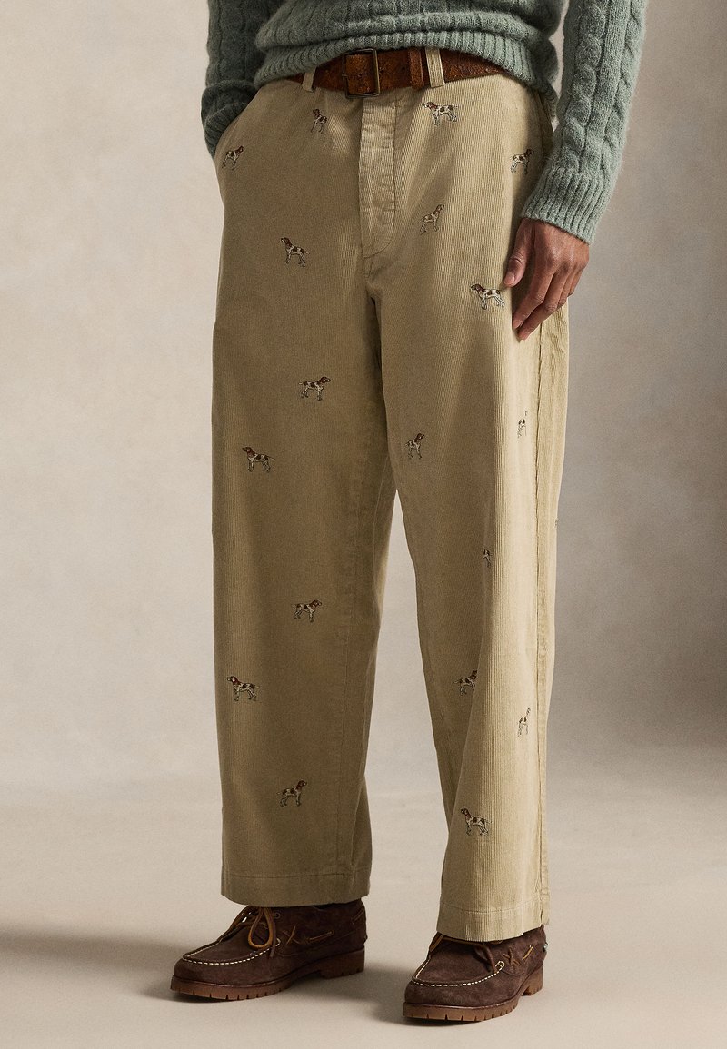 Pantaloni di velluto a coste beige con motivi ricamati di cani; presentano una vestibilità comoda, tessuto strutturato e sono abbinati a mocassini marroni.