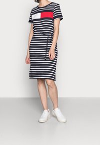 Gestreiftes Kleid in Marineblau und Weiß mit einem roten Brustpaneel, kurzen Ärmeln, Tunnelzug in der Taille und Midi-Länge. Getragen mit weißen Sneakers.