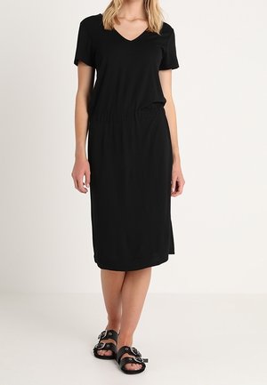 Kobieta w czarnej sukience midi z krótkim rękawem i dekoltem w szpic, z elastyczną talią oraz czarnych sandałach z klamrą, stojąca na tle jasnej, gładkiej powierzchni.