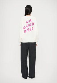 Pletený biely sveter s nápisom "OR GOOD BYES" v jasne ružovej na zadnej strane; má rebrovanú textúru a voľný strih, spárovaný s tmavými nohavicami.