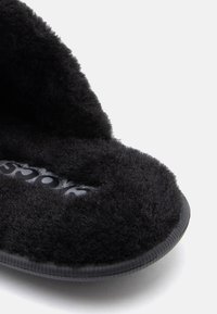 Papuci negri cu un interior textil pufos, formă rotundă și talpă moale. Logo-ul embosat pe interiorul papucului adaugă detaliu.