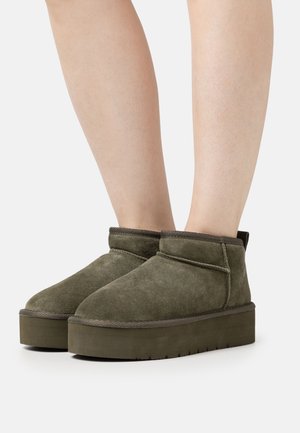 Pieds portant des bottines chevilles en daim vert olive avec des semelles compensées épaisses sur un fond clair.