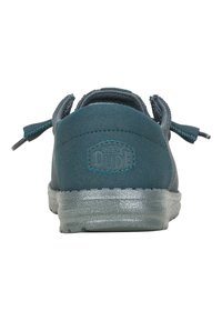 Zapato slip-on de ante en color azul verdoso con una suela texturizada y transparente. La vista trasera presenta dos tiradores y un logo en relieve.