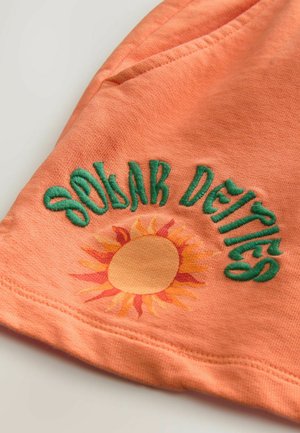 Oranje stof met groene geborduurde tekst "Zonnegoden" en een zonafbeelding met gele stralen. Gladde textuur met gestikte randen.