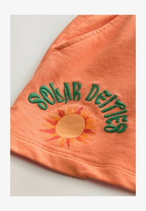 Tissu orange avec le texte brodé en vert "Deités Solaires" et un graphique de soleil avec des rayons jaunes. Texture lisse avec des bords surpiqués.