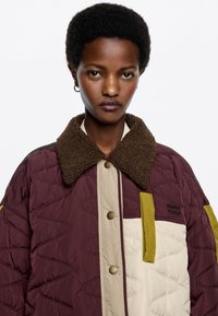 Chaqueta acolchada con un cuerpo color burdeos, panel frontal beige y detalles en verde. El cuello está hecho de un material de felpa marrón, con botones de presión.