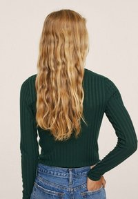 Dunkelgrünes, geripptes Langarmoberteil mit figurbetontem Design, kombiniert mit hochtaillierten blauen Damenjeans. Die Haare sind wellig und fallen über den Rücken.