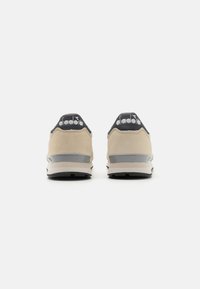 Beige suede athletic shoes with a black logo at the heel, grey accents, and a textured black sole. Design features a low-profile silhouette.