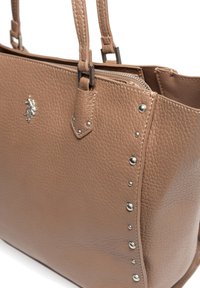 Brązowa skórzana torba tote o teksturowanej powierzchni, z metalowymi akcentami w kolorze srebra i ozdobnymi ćwiekami po jednej stronie. Zamek błyskawiczny na górze.