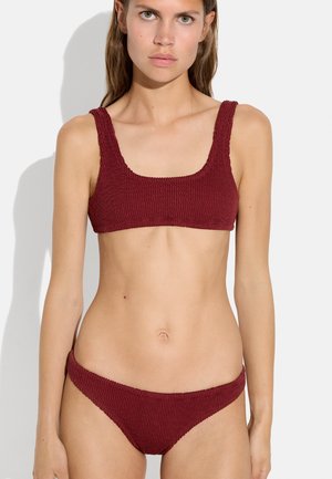 Kobieta ubrana w teksturowany ciemnoczerwony bikini z szerokimi ramiączkami i majtkami z niskim stanem, stoi na prostym jasnym tle.