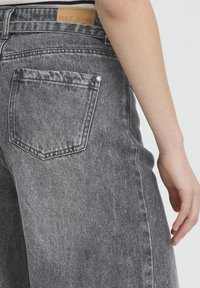 Gros plan sur la poche arrière d'un jean en denim gris avec une étiquette brune PULZ JEANS et le bras d'une personne reposant à côté de la hanche sur un fond uni.