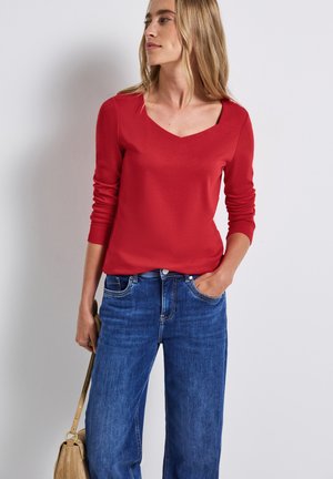 Rotes Langarmoberteil mit V-Ausschnitt, kombiniert mit weiten blauen Jeans aus Denim. Der Stoff wirkt glatt und leicht, geeignet für legere Kleidung.