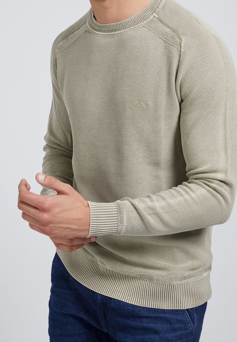 Pull en maille vert olive clair avec un design texturé, des poignets côtelés et un col rond, porté avec un jean bleu.