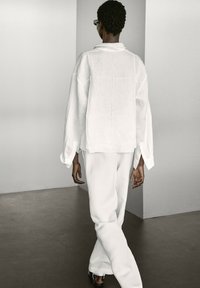 Chemise en lin blanche avec des manches longues et larges, et des fentes sur les côtés, associée à un pantalon blanc ample. Texture lisse, design minimaliste, sans motifs.