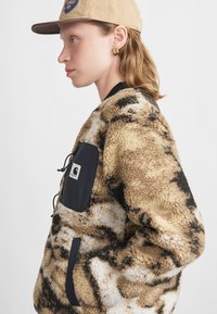 Chaqueta de pelo sintético en tonos marrón claro y blanco con manchas oscuras, que cuenta con un bolsillo de tela de nailon negro y un cordón ajustable, finalizada con un cuello acanalado.