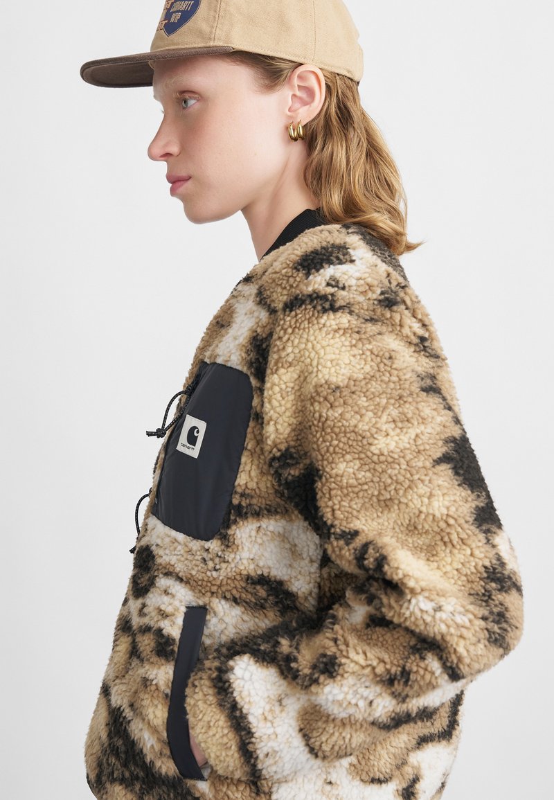 Chaqueta de pelo sintético en tonos marrón claro y blanco con manchas oscuras, que cuenta con un bolsillo de tela de nailon negro y un cordón ajustable, finalizada con un cuello acanalado.