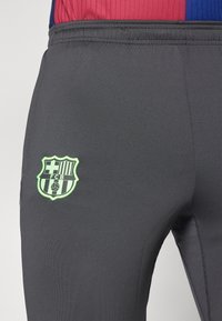 Grijze sportbroek met een groen geborduurd FC Barcelona-logo aan de linkerkant, gemaakt van een gladde, vochtafvoerende stof.
