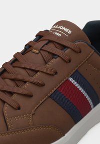 Καφέ αθλητικό παπούτσι με κόκκινη, ναυτική και λευκή ρίγα στο πλάι, καφέ κορδόνια, και την επιγραφή "Jack & Jones Est. 1990" στη γλώσσα.