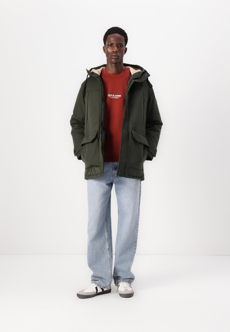 Veste parka verte avec plusieurs poches à l'avant, doublure en polaire beige, associée à un sweat-shirt rouge, un jean bleu clair et des chaussures blanches.