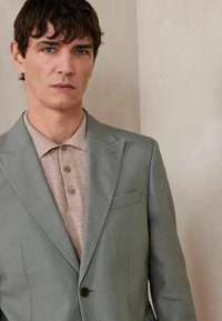 Homme aux cheveux courts et foncés portant un blazer gris clair sur une chemise beige boutonnée, debout contre un mur uni de couleur claire.