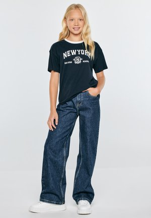 Blondynka stoi z jedną ręką w kieszeni, ma na sobie czarną koszulkę z napisem New York, szerokie ciemnoniebieskie jeansy i białe trampki na jednolitym tle.