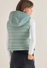 Hellgrüner, gepolsterter Weste mit Kapuze, die horizontale Steppung, Seitentaschen und einen Reißverschluss hat, getragen über einem weißen Langarmoberteil.