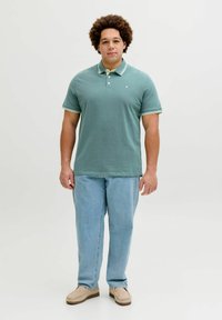 Πόλο πουκάμισο σε τιρκουάζ με γιακά με ρίγες σε λευκό και πράσινο, συνδυασμένο με ανοιχτό μπλε τζιν και μπεζ παπούτσια slip-on, ποζάροντας μπροστά σε ουδέτερο φόντο.