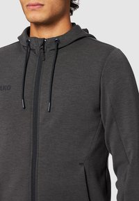 Sweat à zip gris en tissu léger, avec une capuche à cordon et deux poches latérales. Logo discret sur la poitrine.