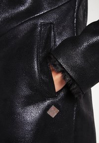 Schwarzer Lederanorak mit einer sichtbaren Hand in einer Seiten tasche, mit einer Fellverzierung am Ärmel und einem silbernen Logoakzent.