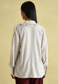 Helle karierte Bluse mit Rüschenkragen, langen Puffärmeln und elastischen Bündchen. Aus weichem Stoff in Beige mit buntem Karomuster.