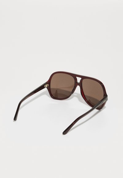 Sunglasses with burgundy plastic frames and brown lenses. Features a distinctive bridge design and sleek, tapered arms with logo accents.