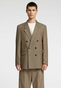 Rjavi dvoredni blazer z velikimi črnimi gumbi, izrezanimi reverji in sprednjimi žepi, kombiniran s svetlimi hlačami. Enobarvno ozadje.