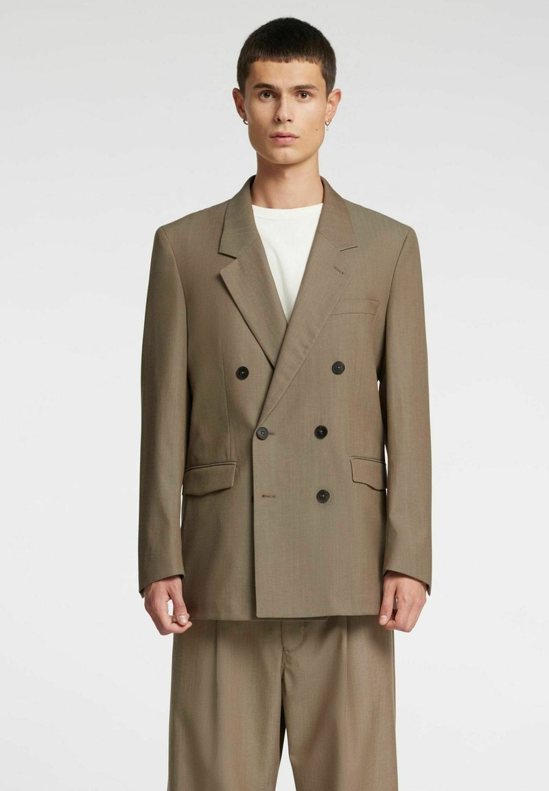 Rjavi dvoredni blazer z velikimi črnimi gumbi, izrezanimi reverji in sprednjimi žepi, kombiniran s svetlimi hlačami. Enobarvno ozadje.