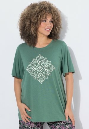 Frau mit lockigem Haar, die ein grünes Kurzarmshirt mit weißem symmetrischem Blumendesign trägt, kombiniert mit gemusterten Hosen.