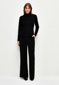 Schwarzes, strukturiertes Langarmoberteil kombiniert mit hochgeschnittenen schwarzen Hosen. Das Modell steht mit einer Hand in der Tasche und trägt spitze schwarze hohe Schuhe.
