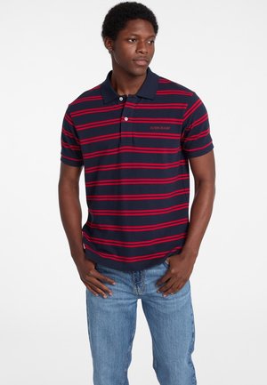 Marineblaue Poloshirt mit roten horizontalen Streifen, zwei-Knopf-Leiste und gesticktem Logo auf der Brust. Kombiniert mit hellblauen Jeans.