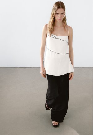 Haut sans manches blanc avec un détail de ligne noire diagonale, associé à un pantalon noir ample. Tissu texturé avec un design fluide.
