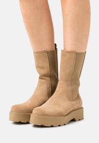 Tan ankle boots made of suede feature a chunky platform sole with a textured finish and exposed stitching on the upper.